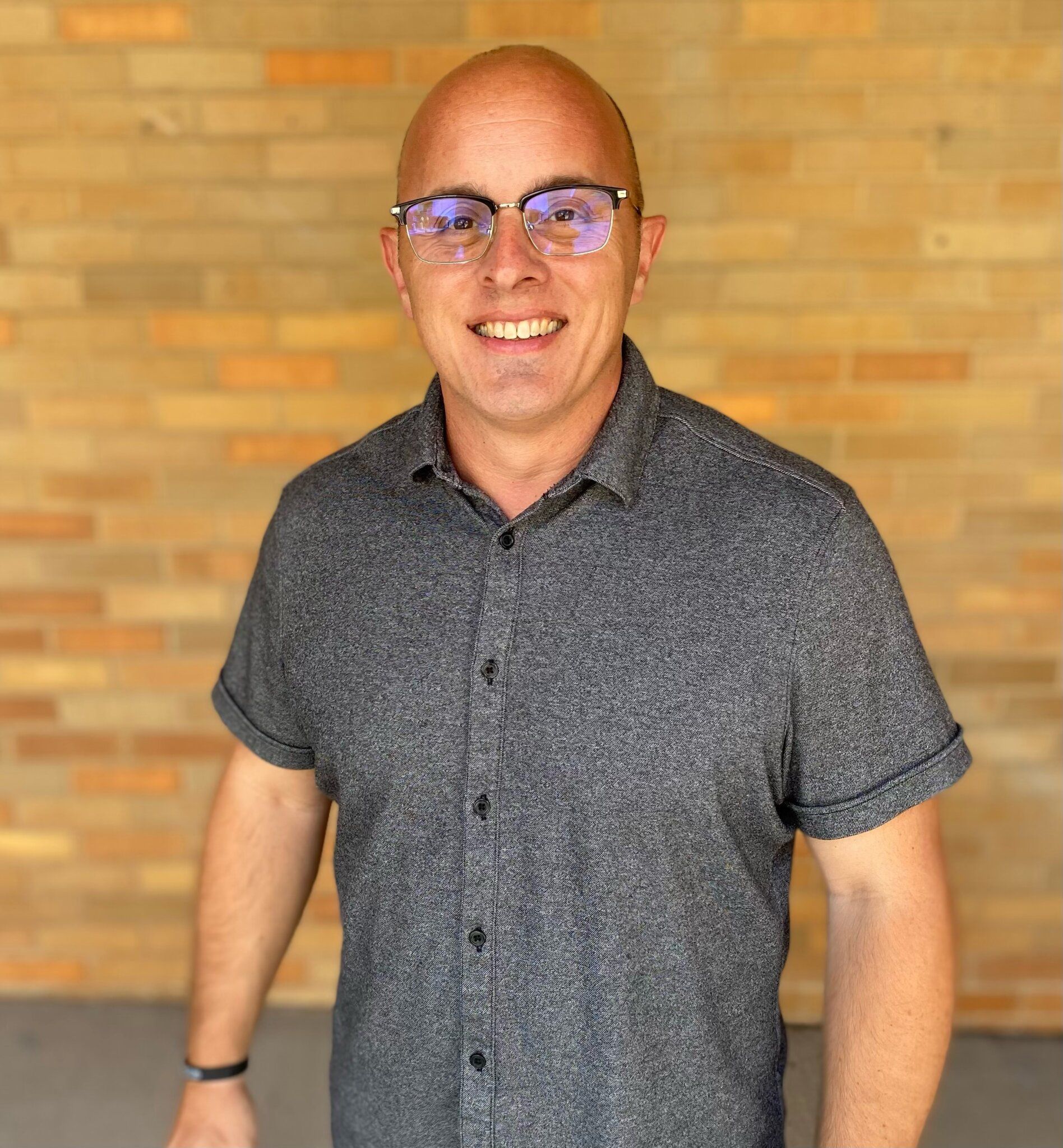 Man in glasses smiles, wearing a gray shirt, in front of a brick wall.