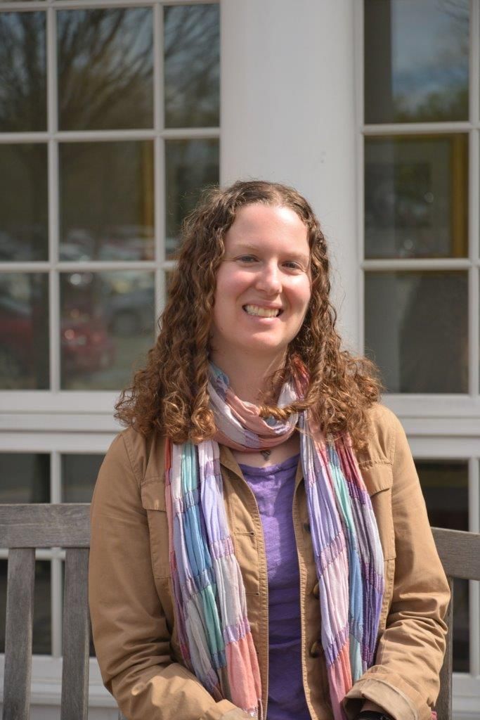 Woman with curly brown hair, smiling, wearing a scarf, jacket, and purple shirt, sitting on a bench in front of a building.