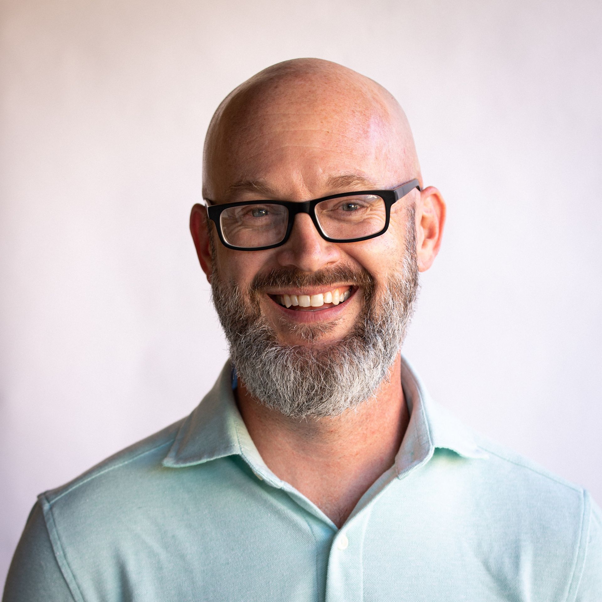 Bald man with a gray beard smiles while wearing glasses and a light blue polo shirt.