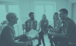 People in a circle, smiling, laughing in a group therapy session. Indoor setting, blue hue.