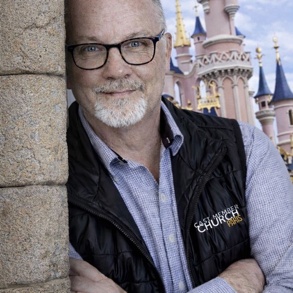 Smiling bald man in a light blue button-up shirt outdoors, with blurred greenery behind him