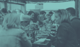 People seated at a table outdoors, eating and conversing; teal-toned.