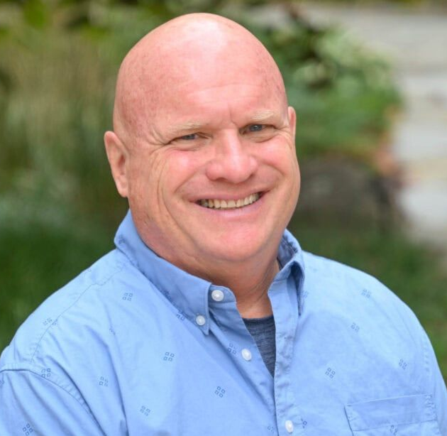 Smiling bald man in a light blue button-up shirt outdoors, with blurred greenery behind him