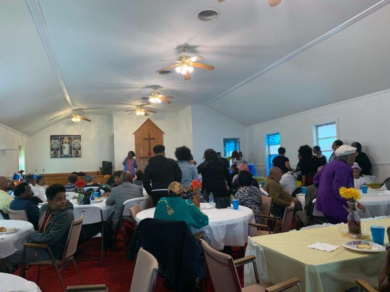 People at tables in a church hall, likely a reception. White tablecloths, red carpet, and ceiling fans.