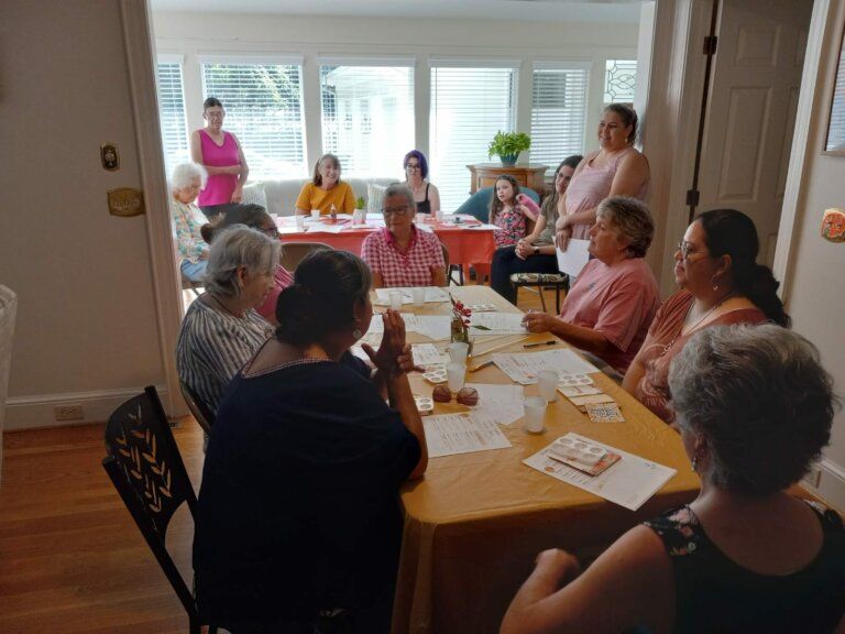 A group of women seated around a table in a bright room. Some are talking, papers present.