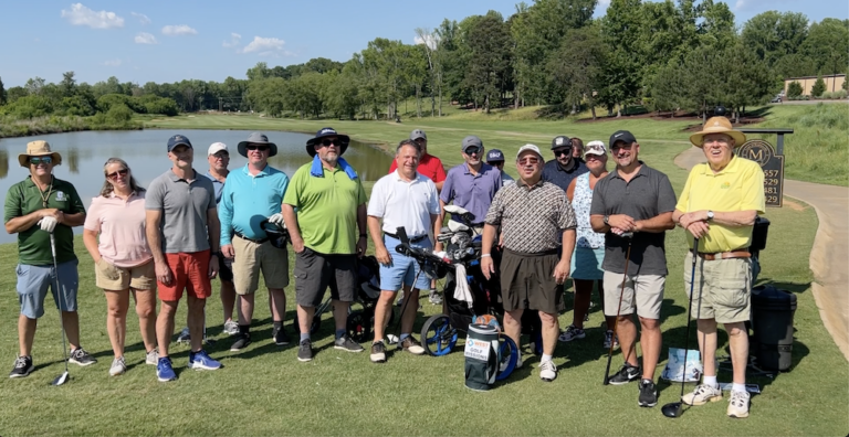 Group of golfers on a course, posing with clubs and bags near a pond on a sunny day.