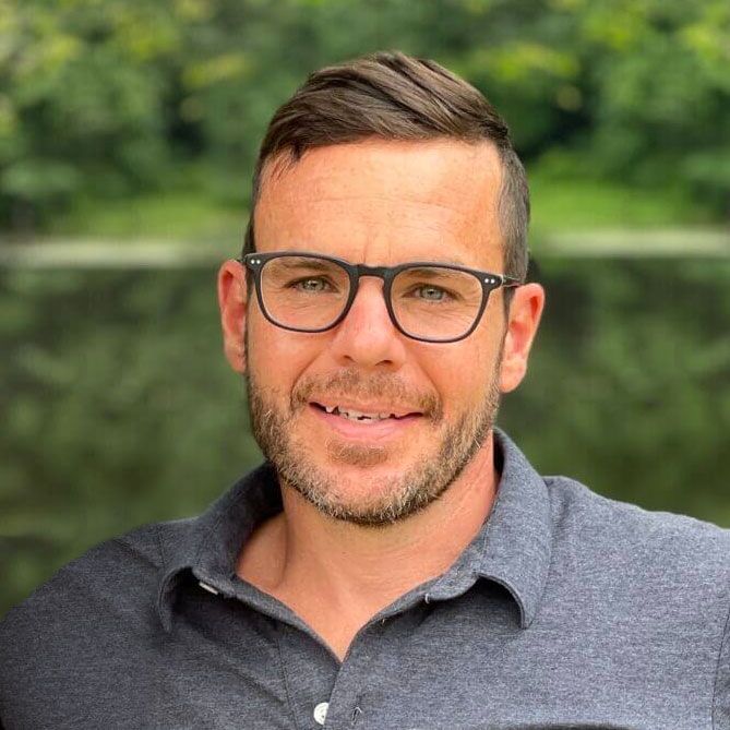 Man with glasses, short brown hair, gray shirt smiles outdoors by water.