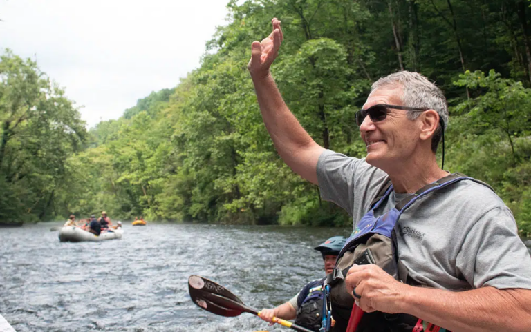 Man waving from a raft on a river, surrounded by green trees.