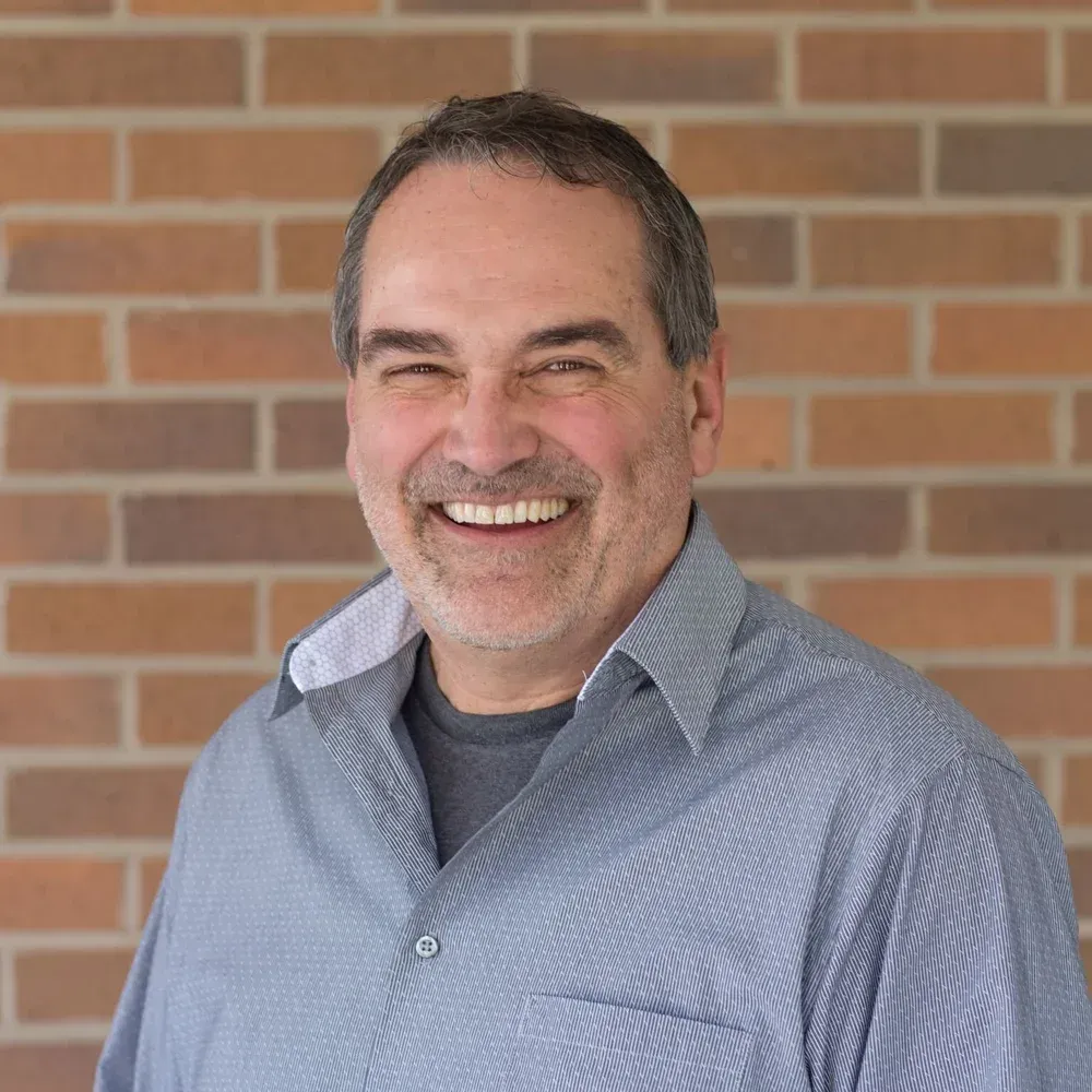 Smiling man in blue shirt stands in front of a brick wall.