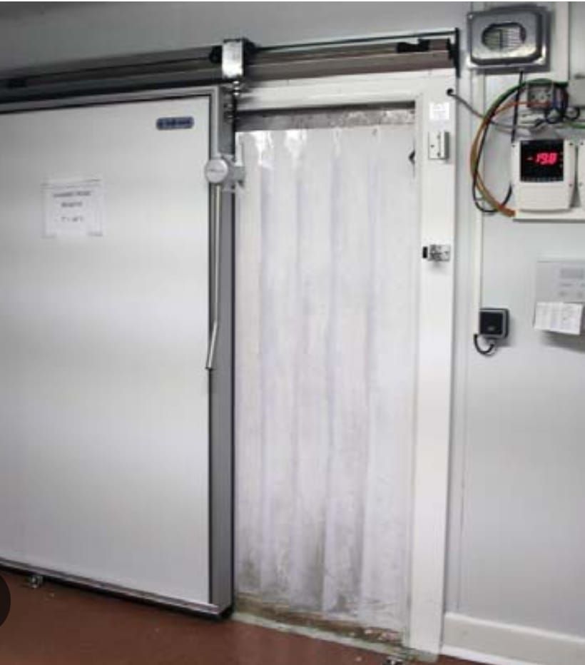 Puerta corrediza de un congelador, abierta con cortinas de plástico transparente. Indicador de temperatura en la pared.
