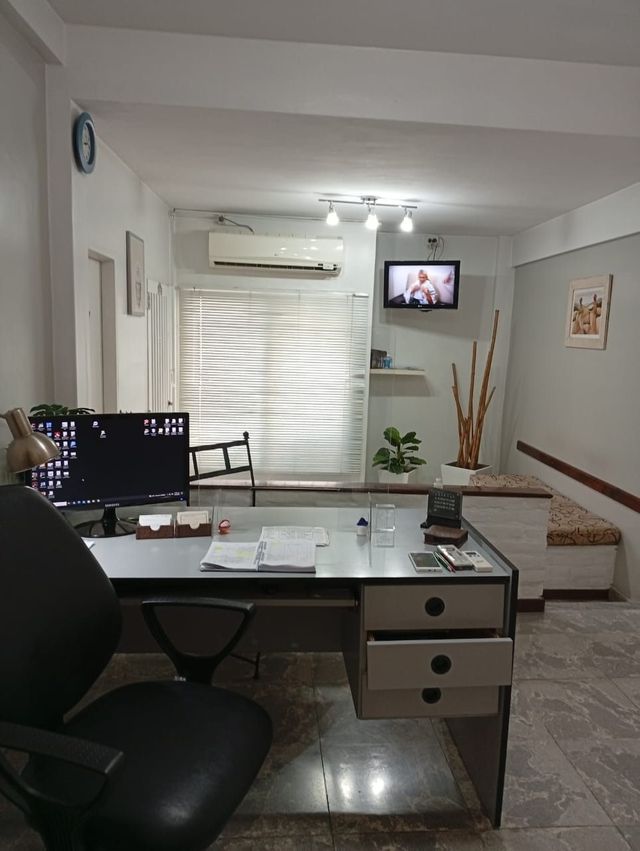 Oficina moderna con escritorio, ordenador, silla, televisor montado en la pared y persianas luminosas.