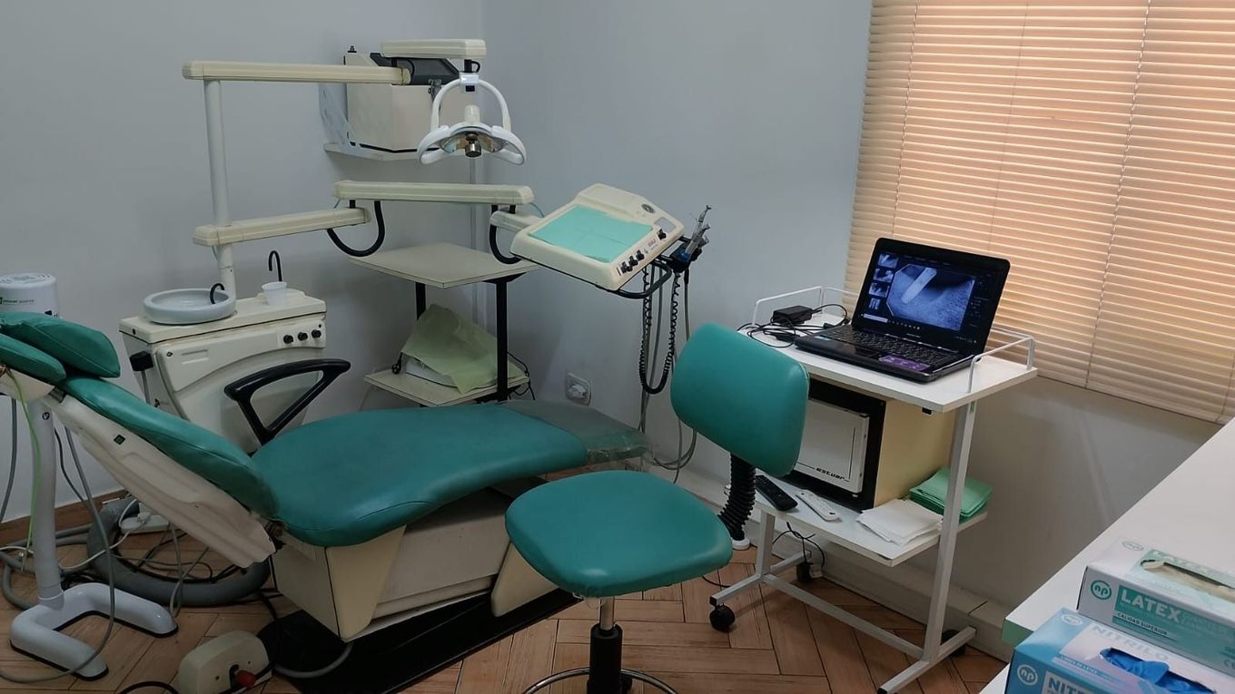 Consultorio dental con sillón color turquesa, equipo médico y una computadora portátil sobre una mesa pequeña.