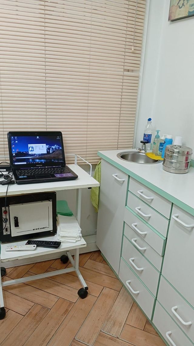 Pequeña sala de exploración con un ordenador portátil en un carrito, armarios y material médico junto a una persiana.