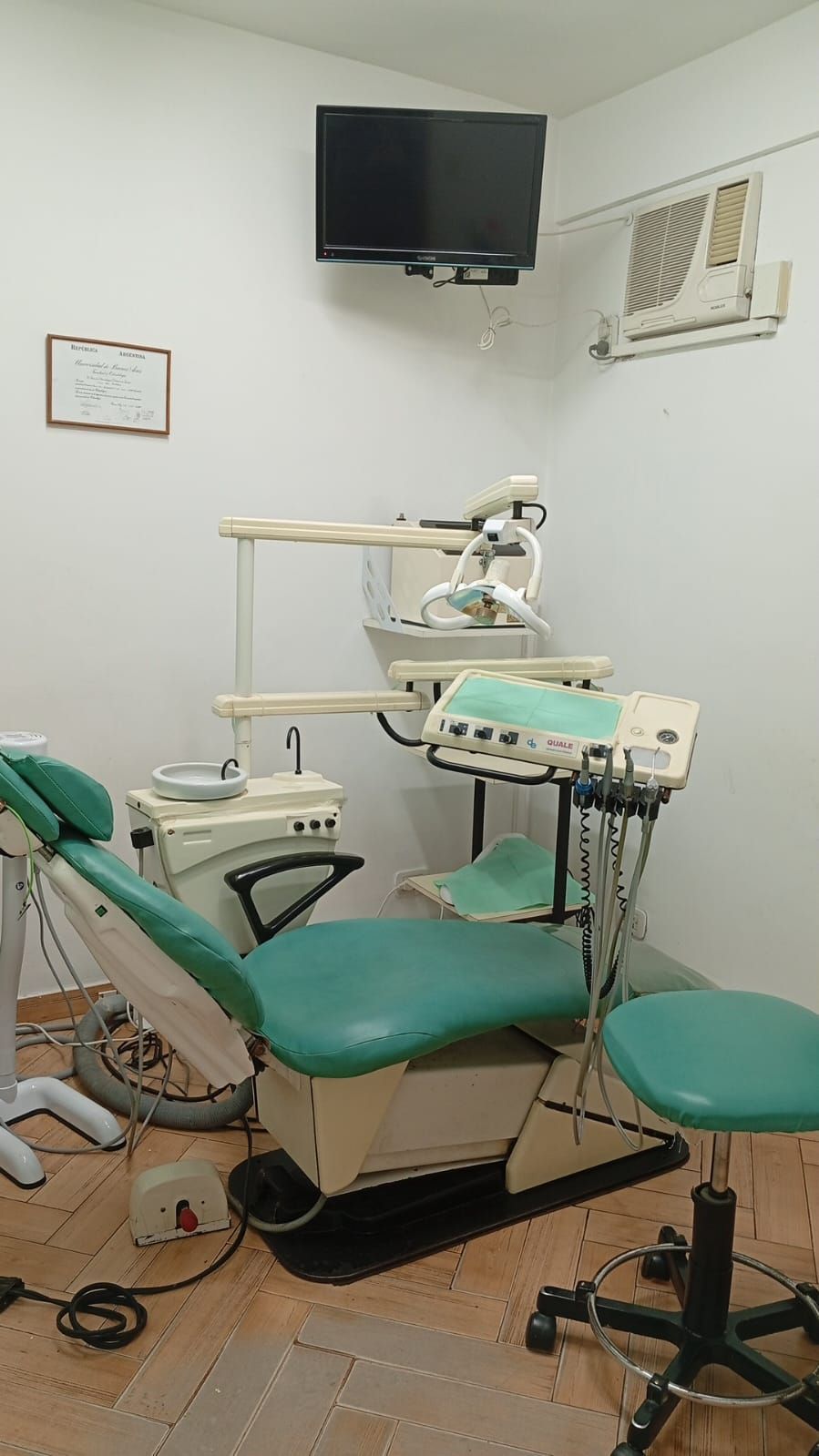 Sala de exploración dental con sillón verde, instrumental dental, monitor de pared y unidad de rayos X superior.