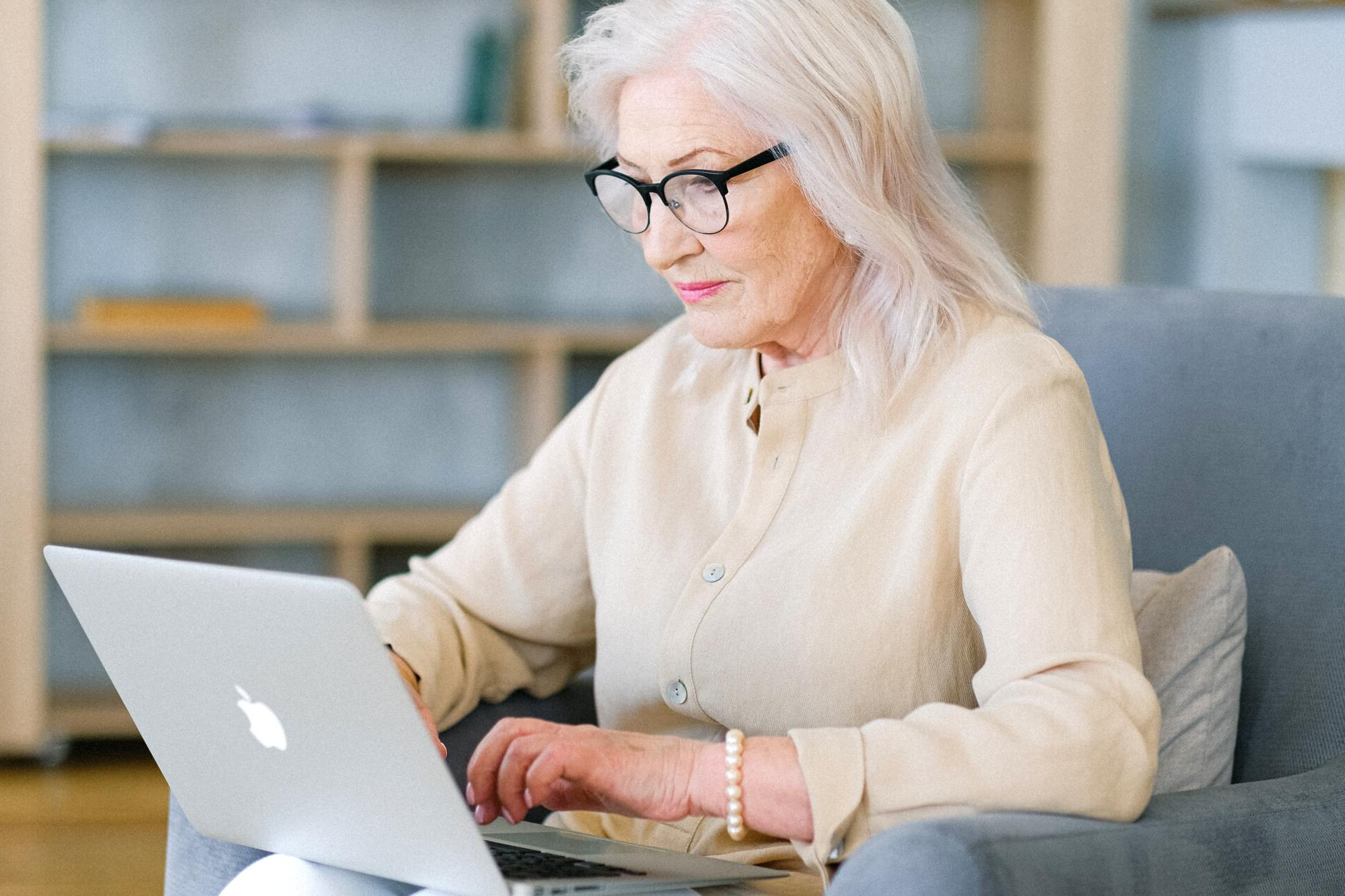 An elderly woman is sitting in a chair using a laptop computer.