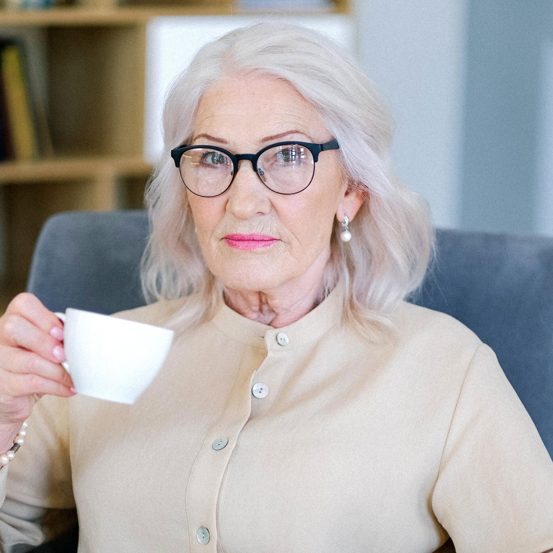 An elderly woman wearing glasses is sitting in a chair holding a cup of coffee.