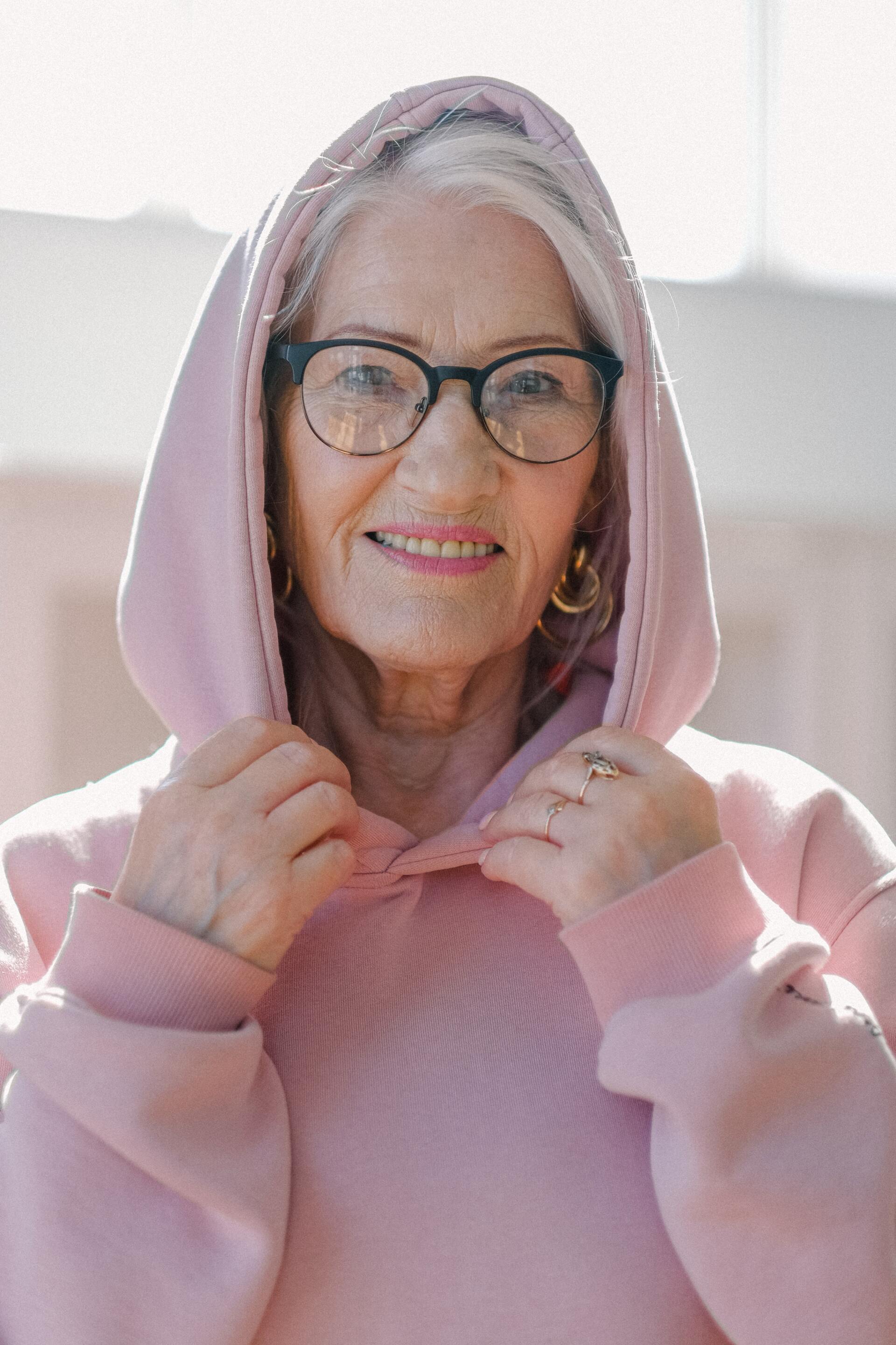 An elderly woman wearing glasses and a pink hoodie is smiling.
