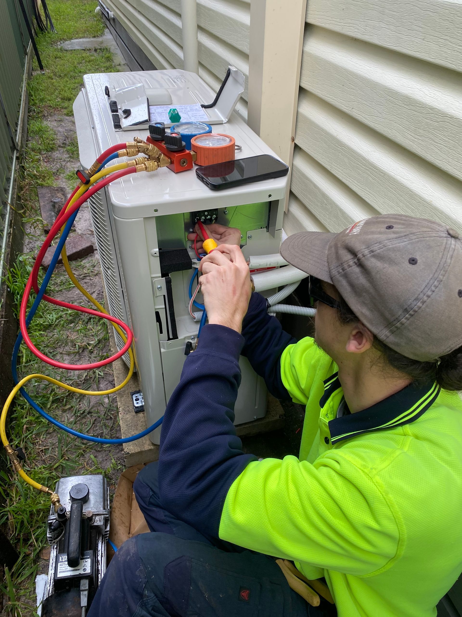 A technician in a safety vest repairs an air conditioning unit outside a building, using tools and gauges. — Airweld Services Pty Ltd in Shellharbour, NSW
