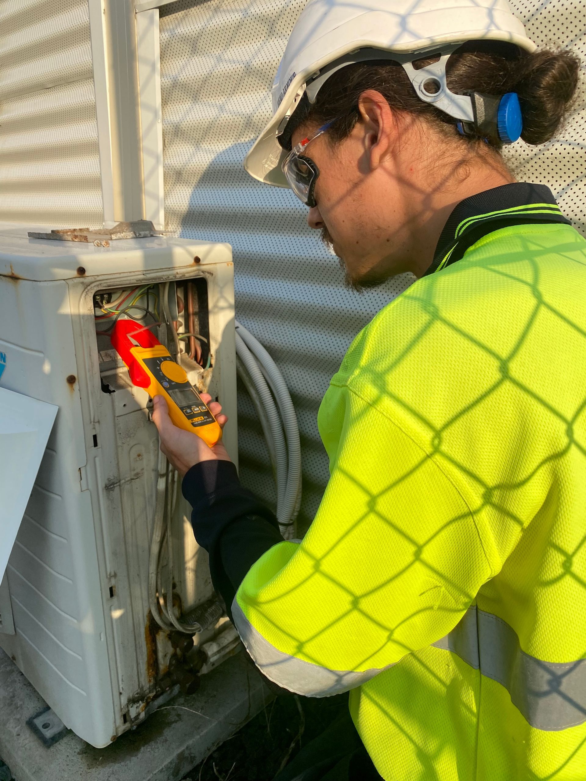 Person in a safety vest inspects AC unit with a yellow device, outdoors near a metal structure. — Airweld Services Pty Ltd in Shellharbour, NSW