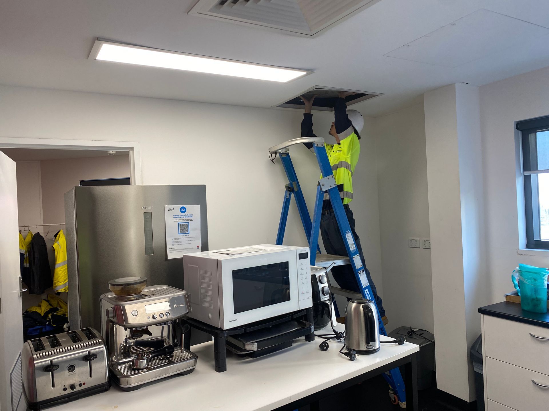 Person on a ladder, working on a ceiling panel in a break room. Includes microwave, fridge, and coffee maker. — Airweld Services Pty Ltd in Kiama, NSW