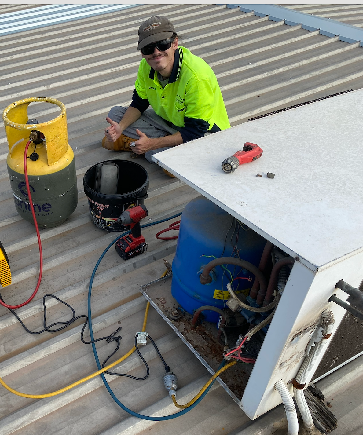HVAC technician on roof, repairing unit. Yellow refrigerant tank, tools, blue component, thumbs up — Airweld Services Pty Ltd in Shellharbour, NSW