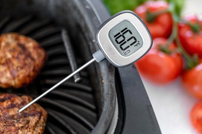 A digital thermometer is sitting on a grill next to a hamburger and tomatoes.