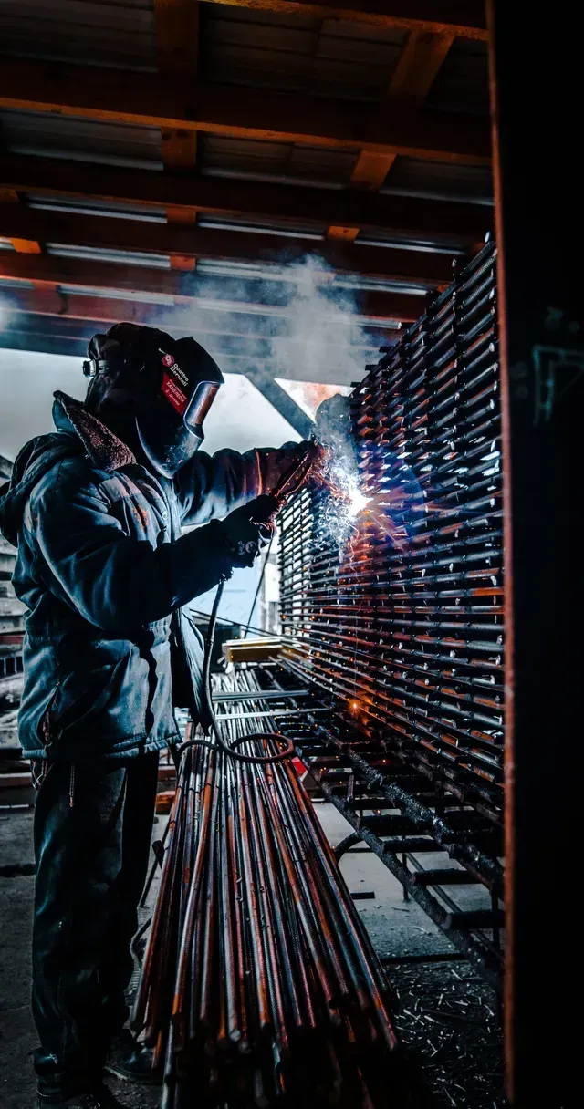 A picture of a man in full welders clothes standing up in a shop and welding rebar at shoulder level with black welders helmet on.  He is using a TIG welder.  There is a stack of rebar on a rack in front of him and he is slightly leaning over it. 