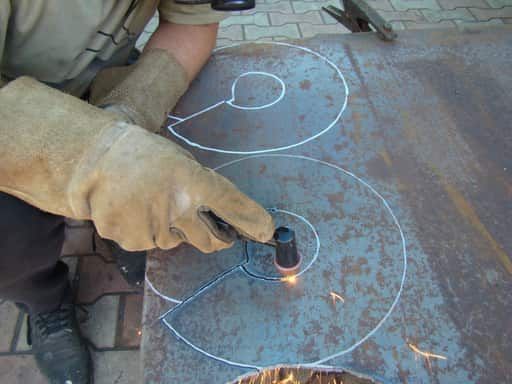 Close up of a person using a hand piece for a plasma cutter to cut sheet steel.  The steel sheet has spiral patterns drawn on it with white chalk.  The person is cutting following the lines.