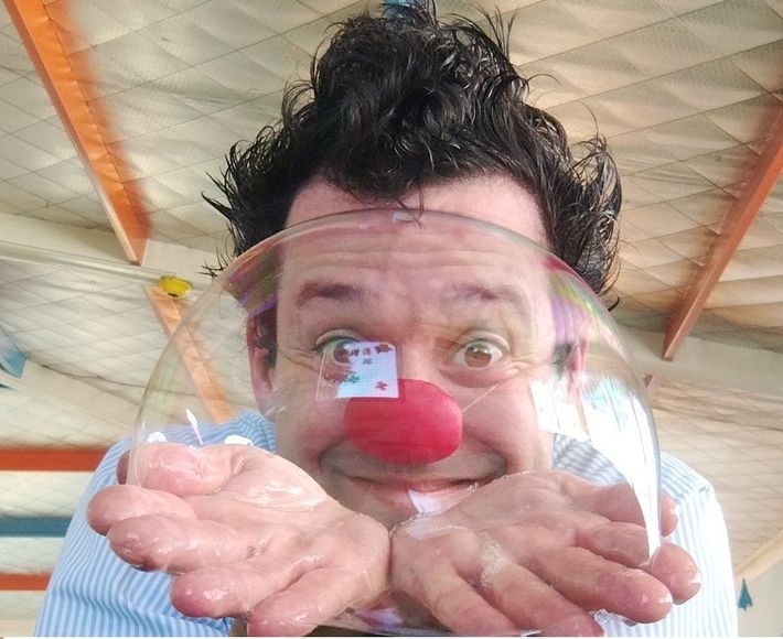 Clown with red nose, inside a large soap bubble, smiling with open palms.