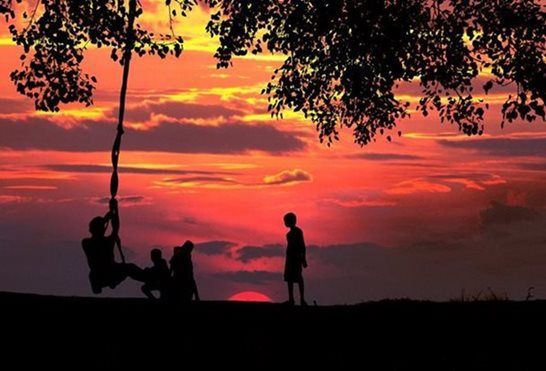 Silhouetted people on a swing under trees at sunset with a vibrant red-orange sky