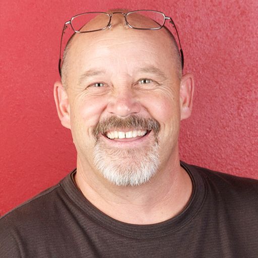 Smiling man with a bald head, glasses on top, and a goatee against a red background.
