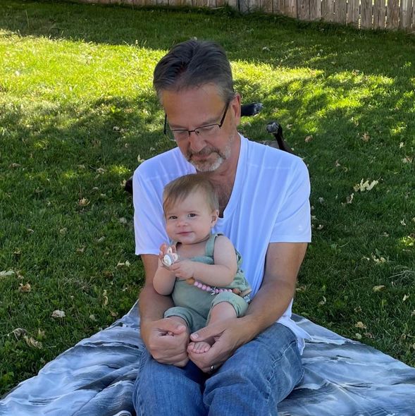 Man sitting outside holding a baby, both on a blanket. Baby has a pacifier.