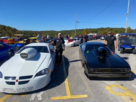 Two drag racing cars lined up on a track, one white and one black, with spectators in the background