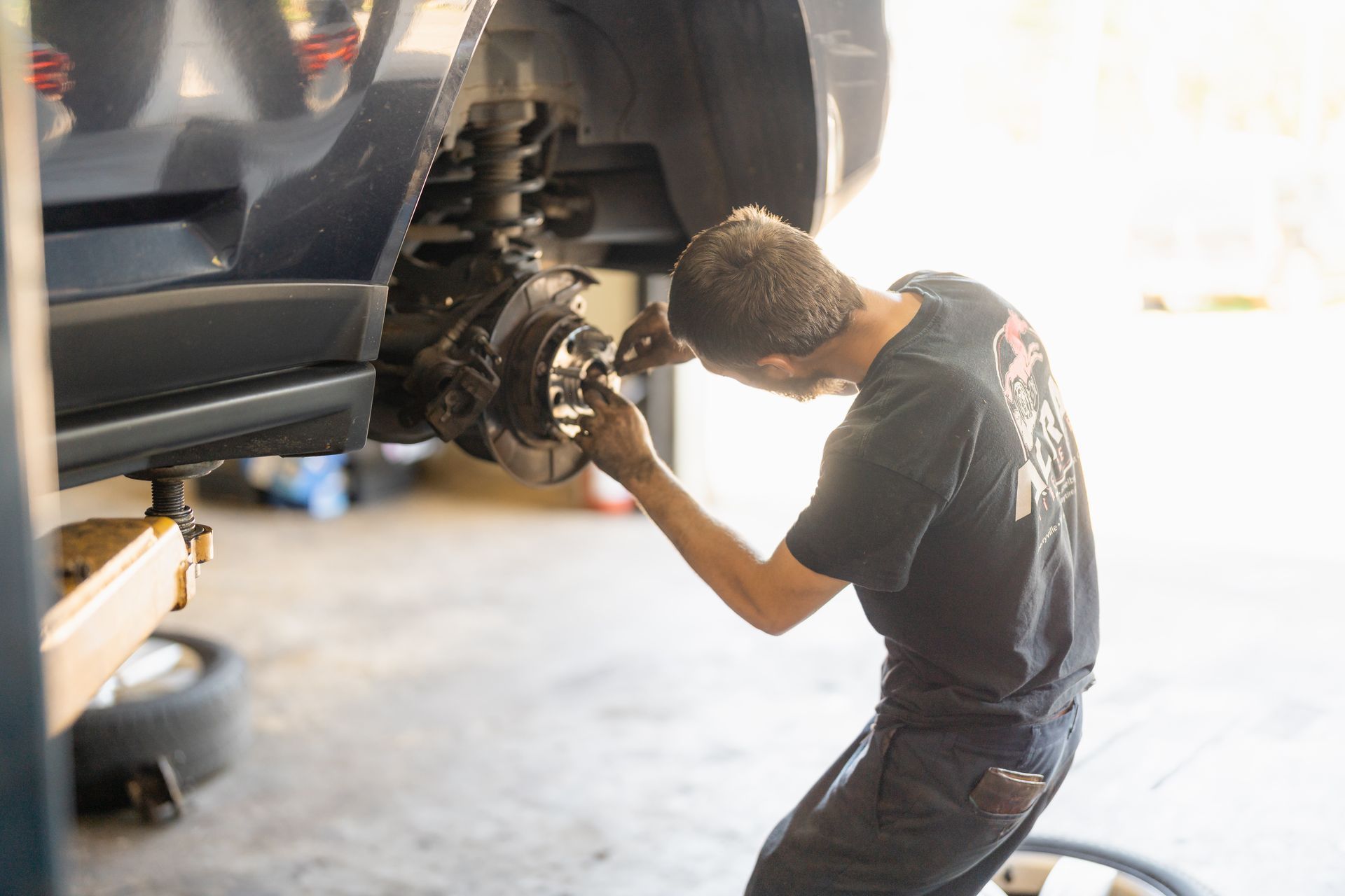 Mechanic repairs a car’s exposed wheel assembly in a garage, bent over under the vehicle.