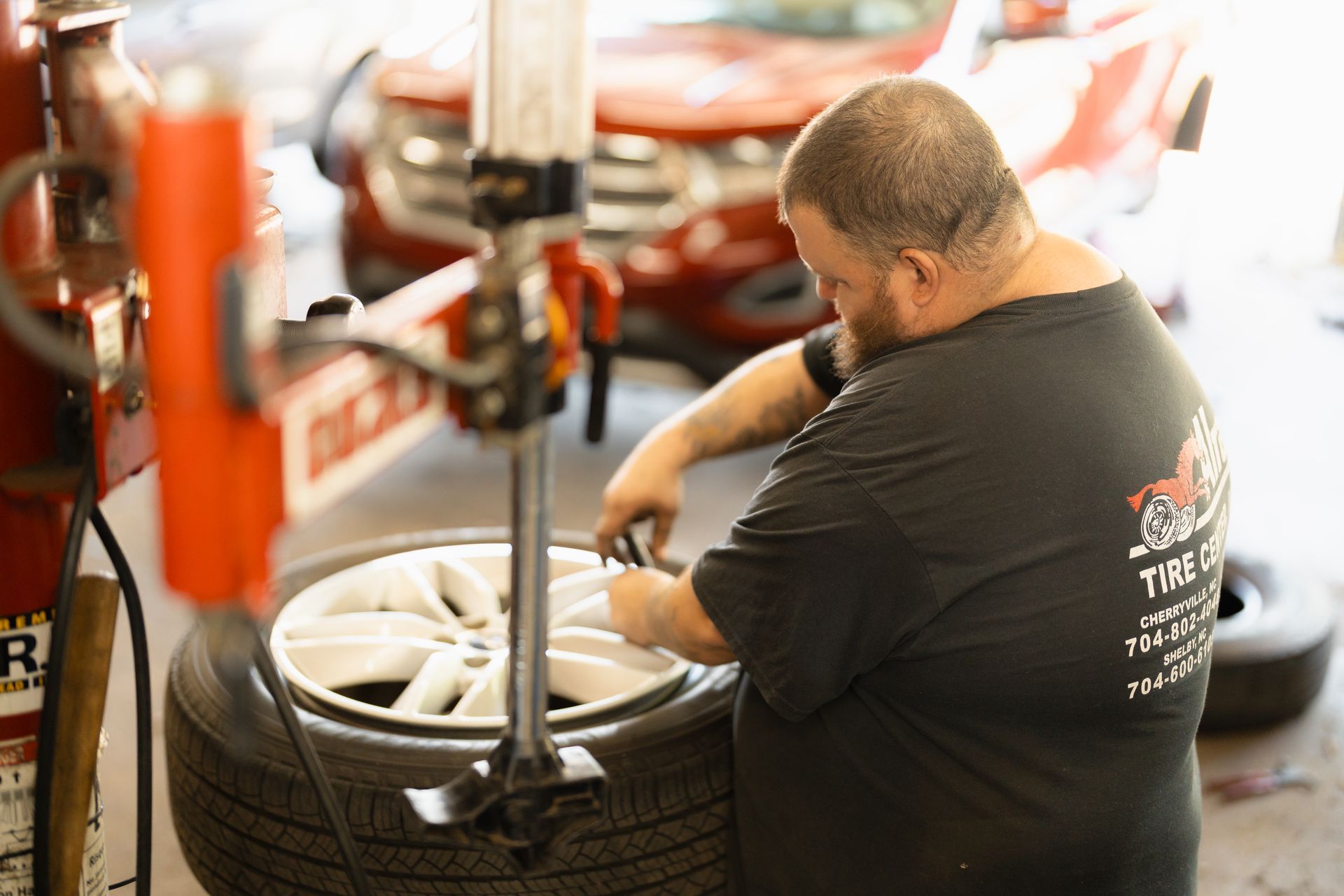 Mechanic crouches beside a tire, using a tool in a shop with red equipment and stacked tires.