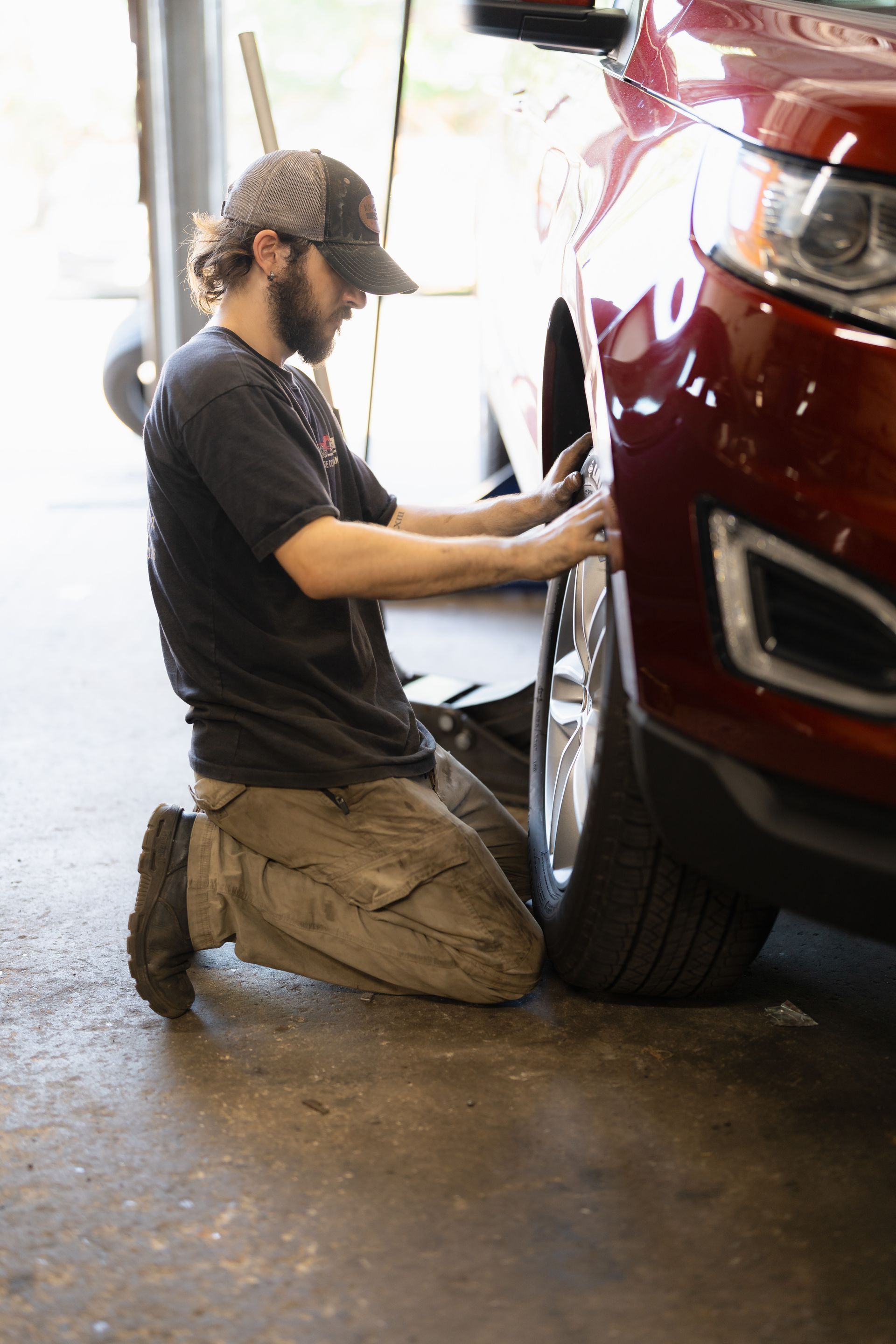 Mechanic kneels beside a red SUV, working on the front wheel in a garage.