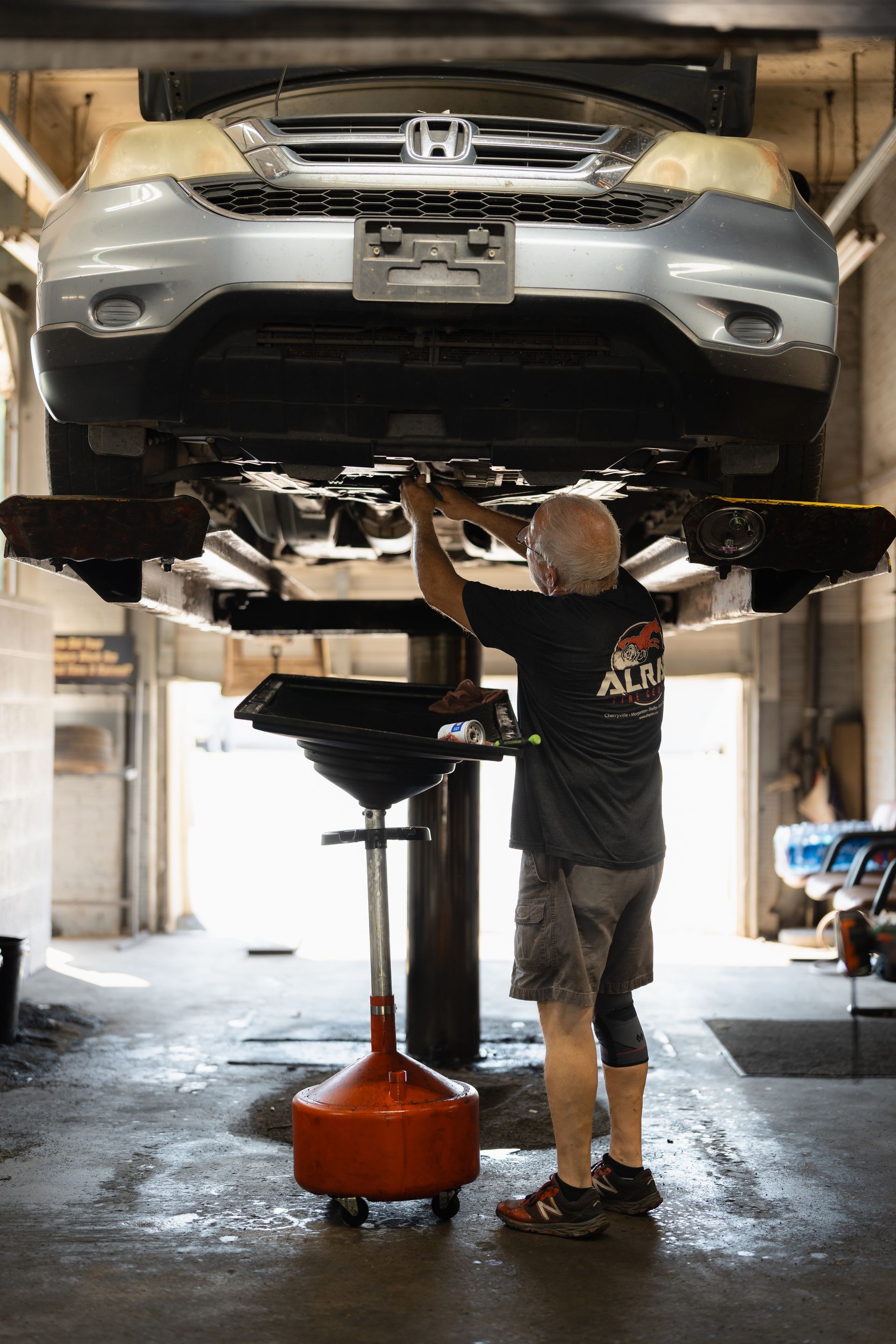 Mechanic draining oil from a silver car lifted on a repair hoist in a garage
