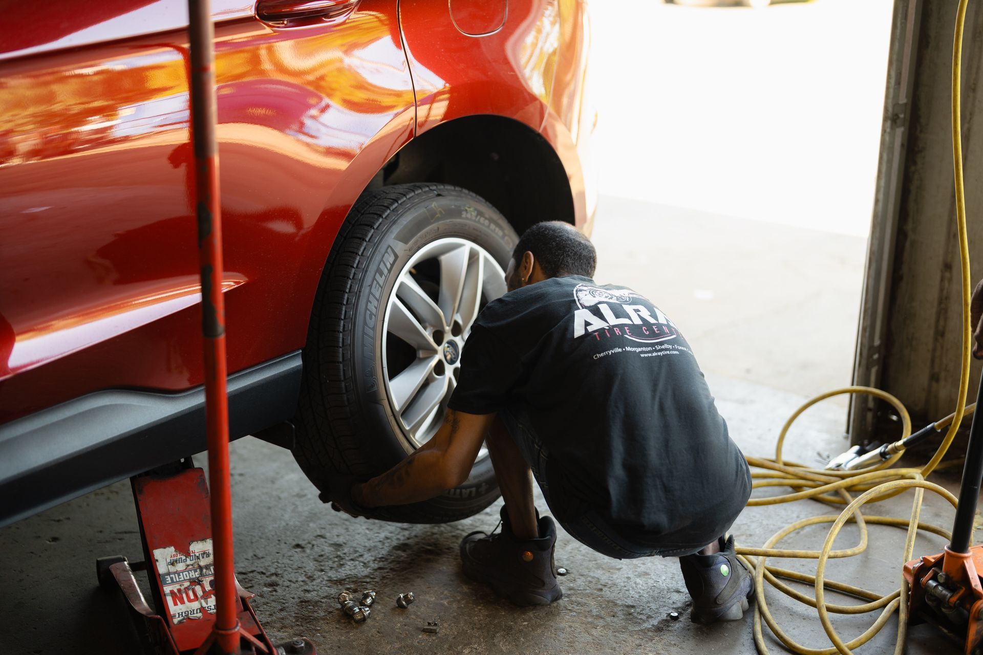 Mechanic crouching by a red car’s rear wheel in a garage, using a tool with yellow cables nearby