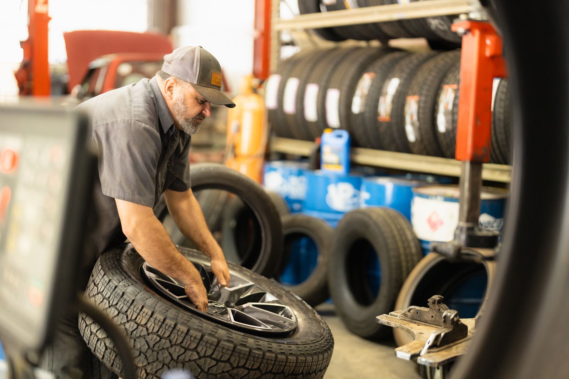 Mechanic fitting a tire onto a wheel in an auto shop, surrounded by stacked tires and tools.