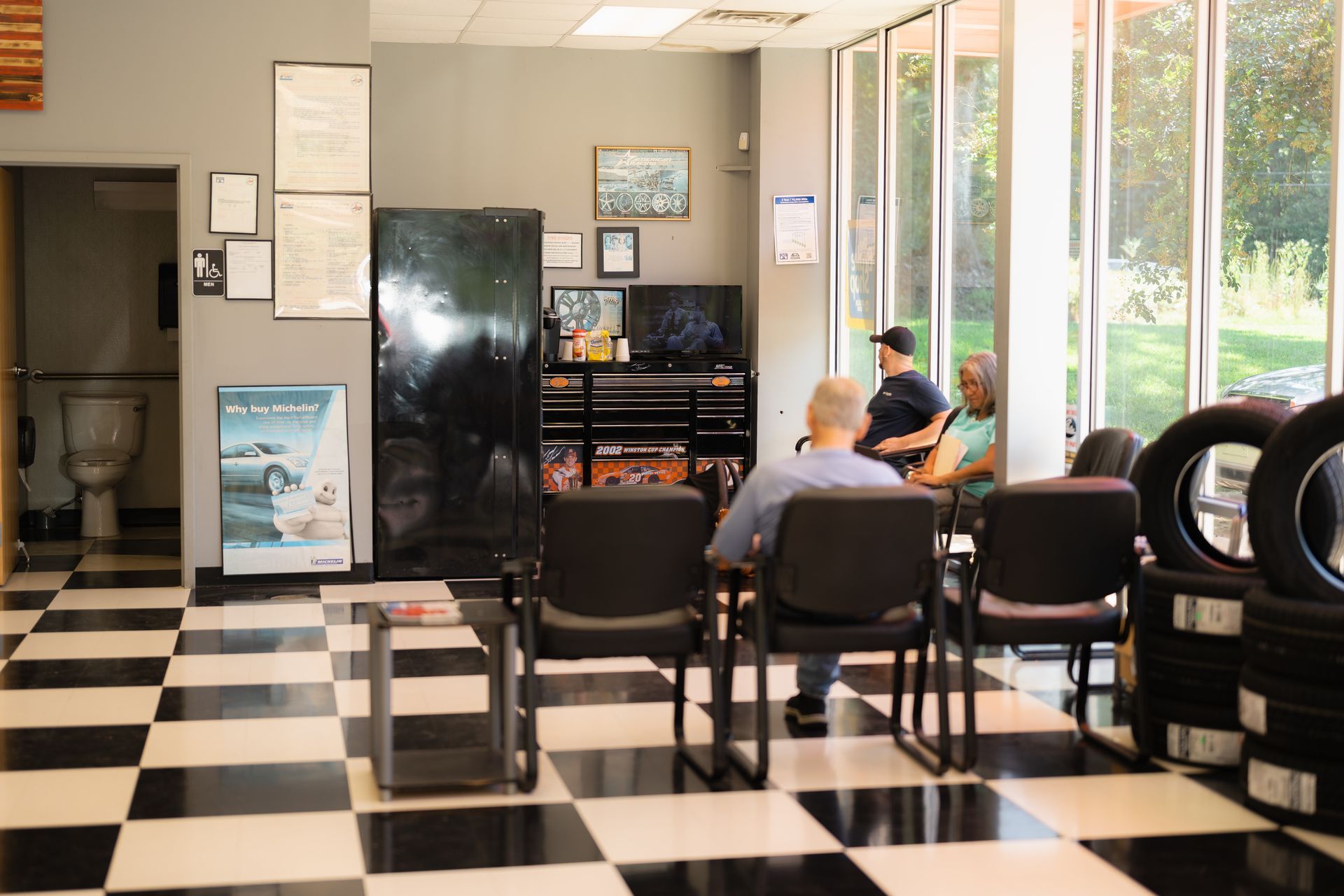 Barbershop waiting area with checkered floor, chairs, wall mirrors, and a person seated by the window