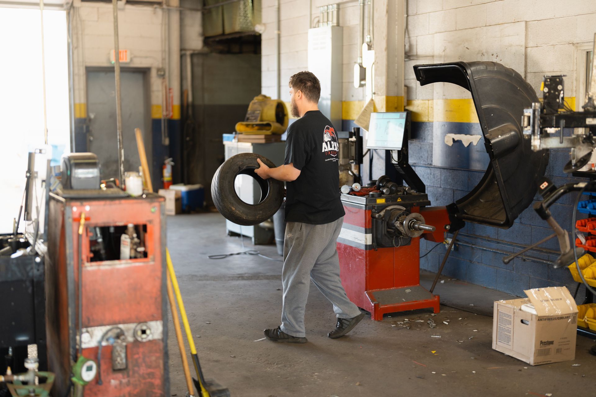 Man in black shirt carrying a tire in an auto repair shop beside machinery and a car lift