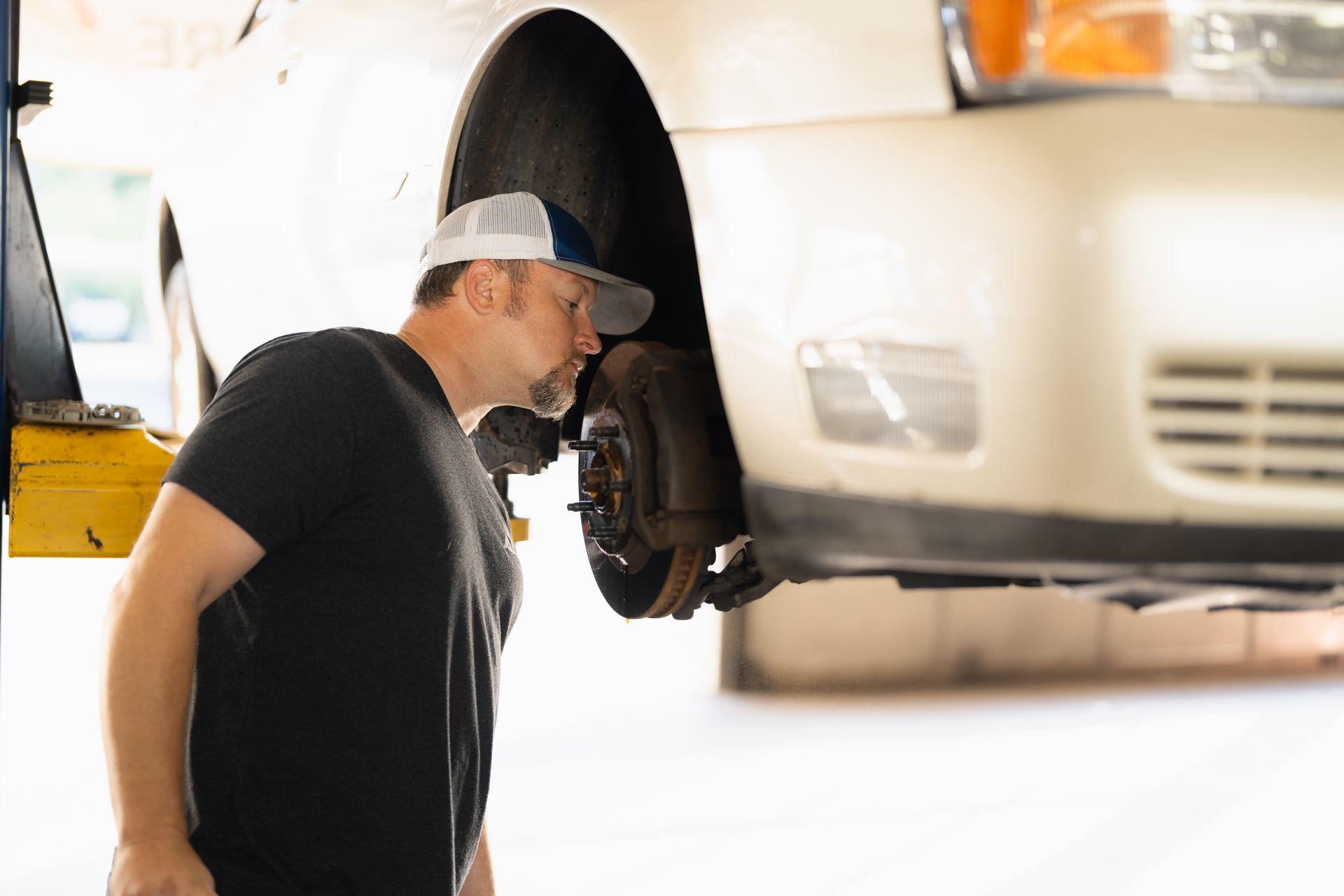 Mechanic inspecting a car’s exposed wheel in a garage, with a white vehicle on a lift nearby
