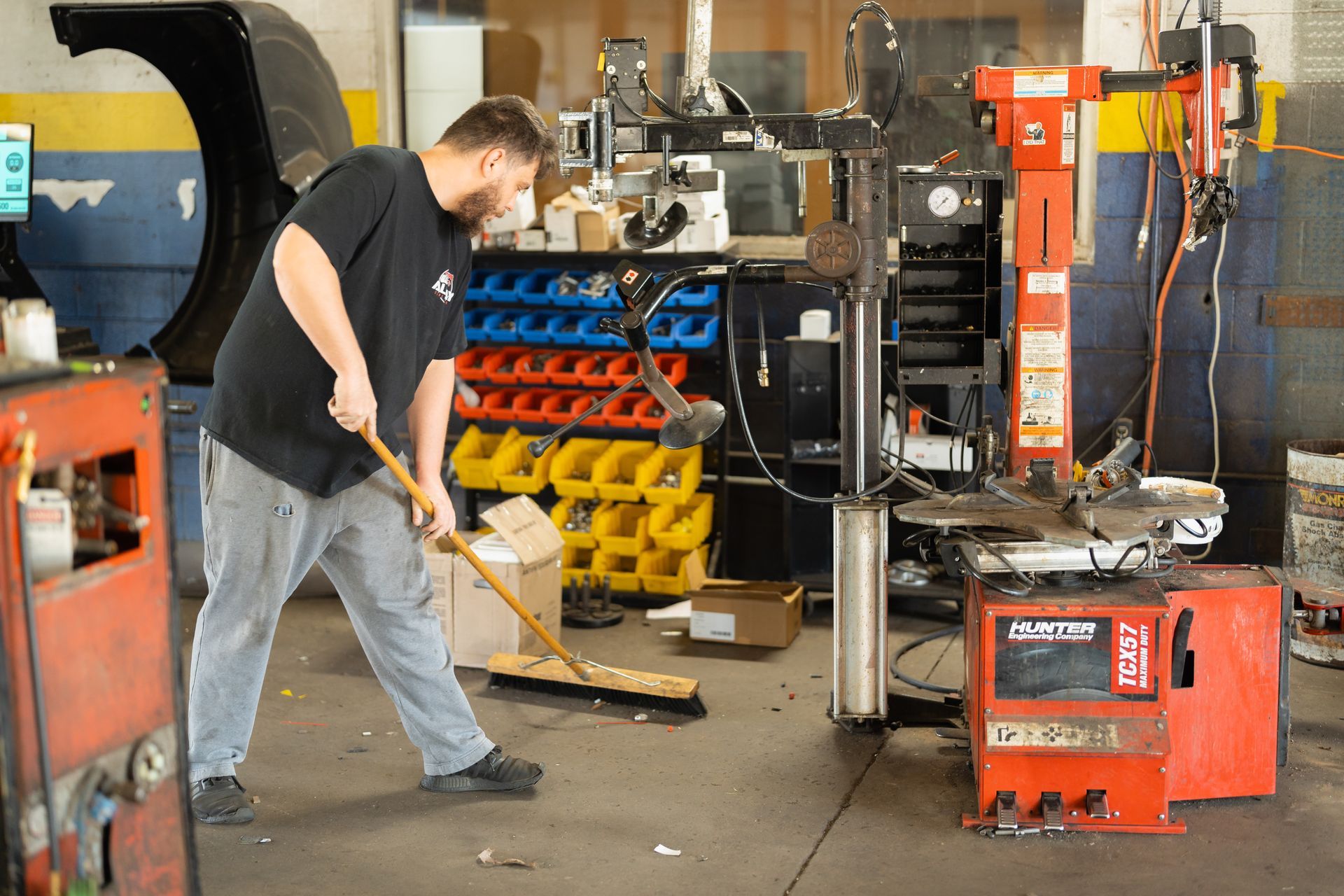 Mechanic sweeping a cluttered auto repair shop beside tools, machinery, and a red welding unit