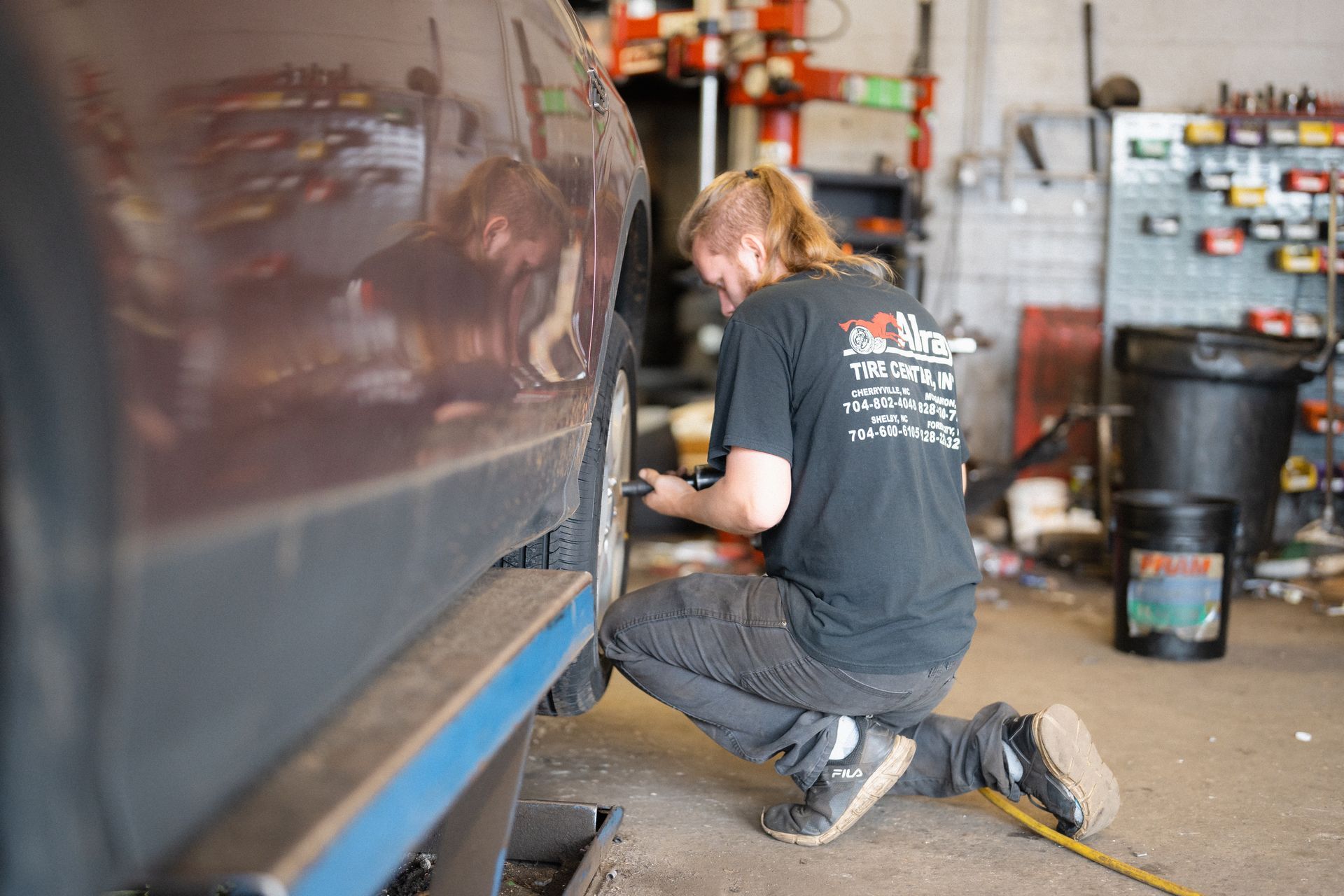 Mechanic kneeling beside a dark car in a garage, working near the wheel well with tools.