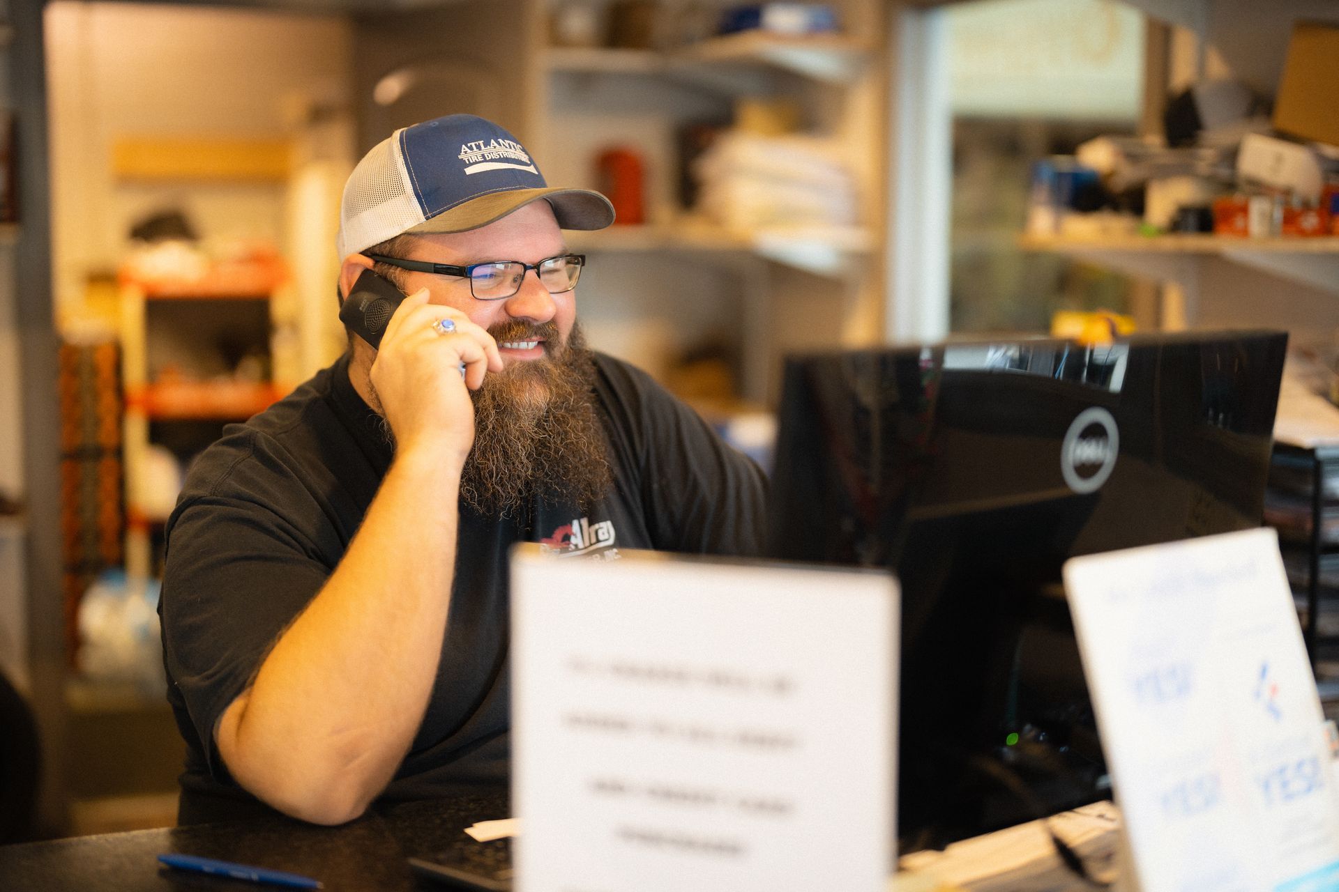 Bearded man on a phone at a desk, working behind a laptop in a warm office setting.
