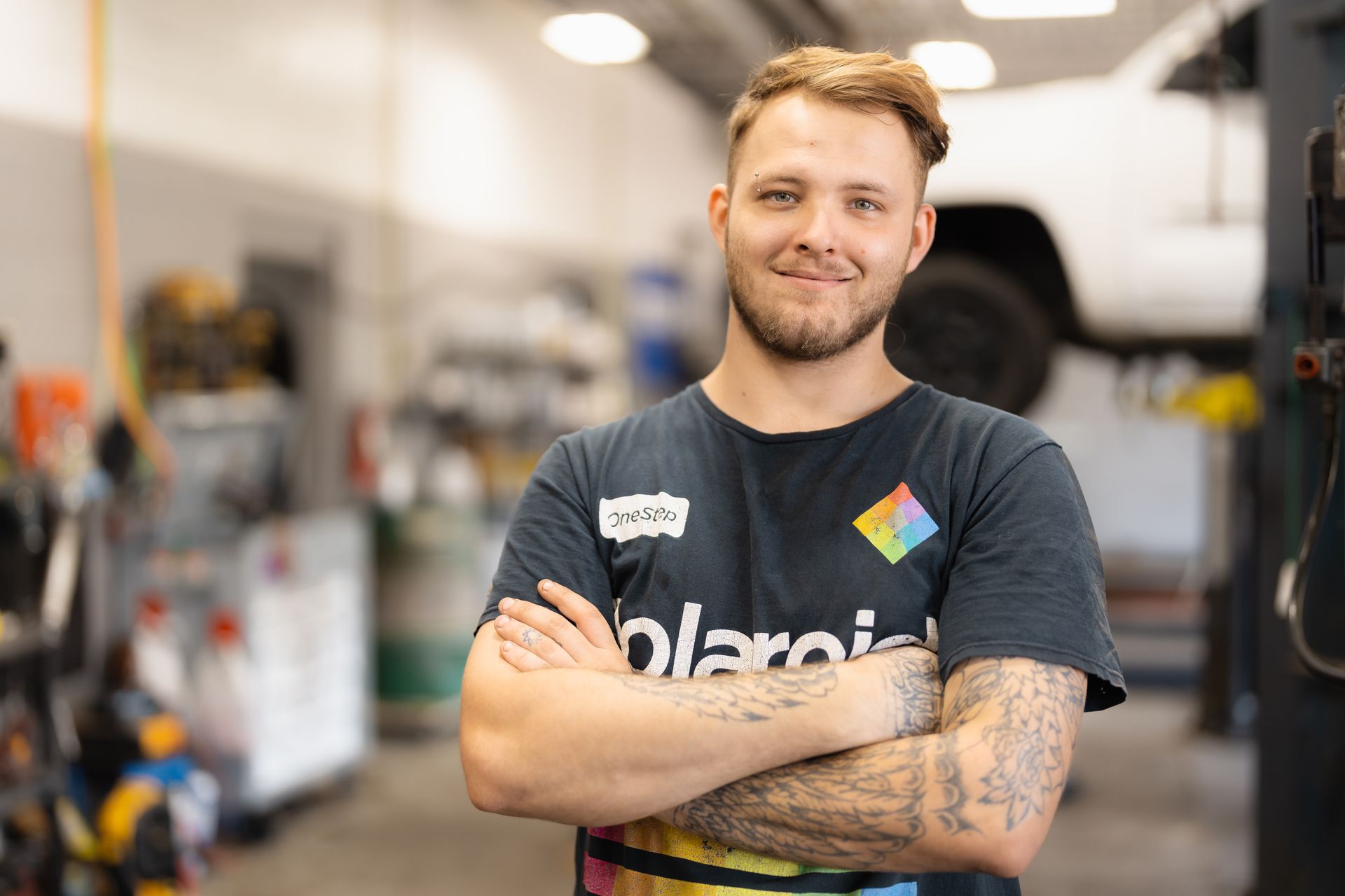 Smiling mechanic with crossed arms in a garage, wearing a dark shirt with tattoos visible.