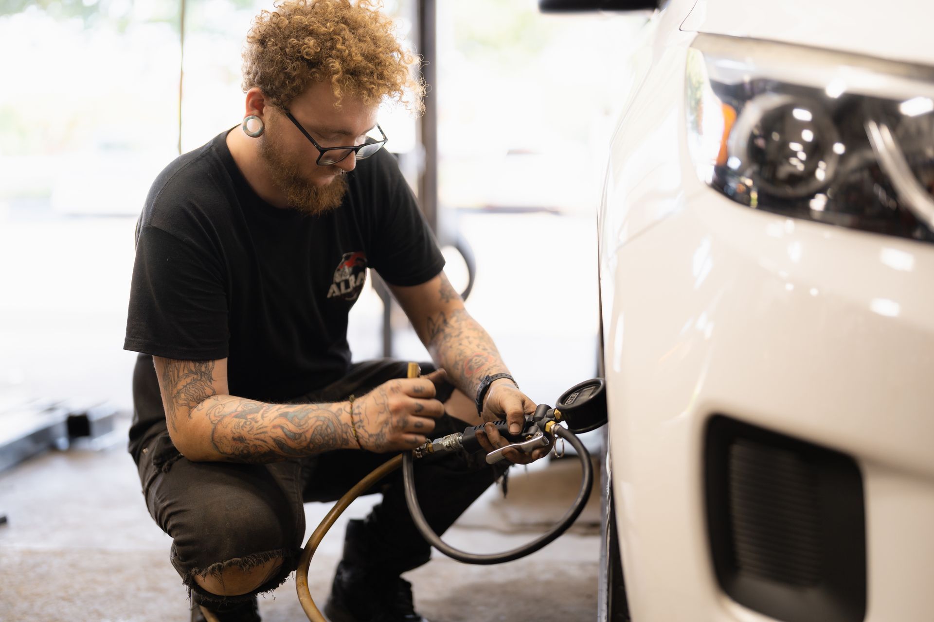Technician kneeling beside a white car, using tools to repair a wheel or brake assembly.