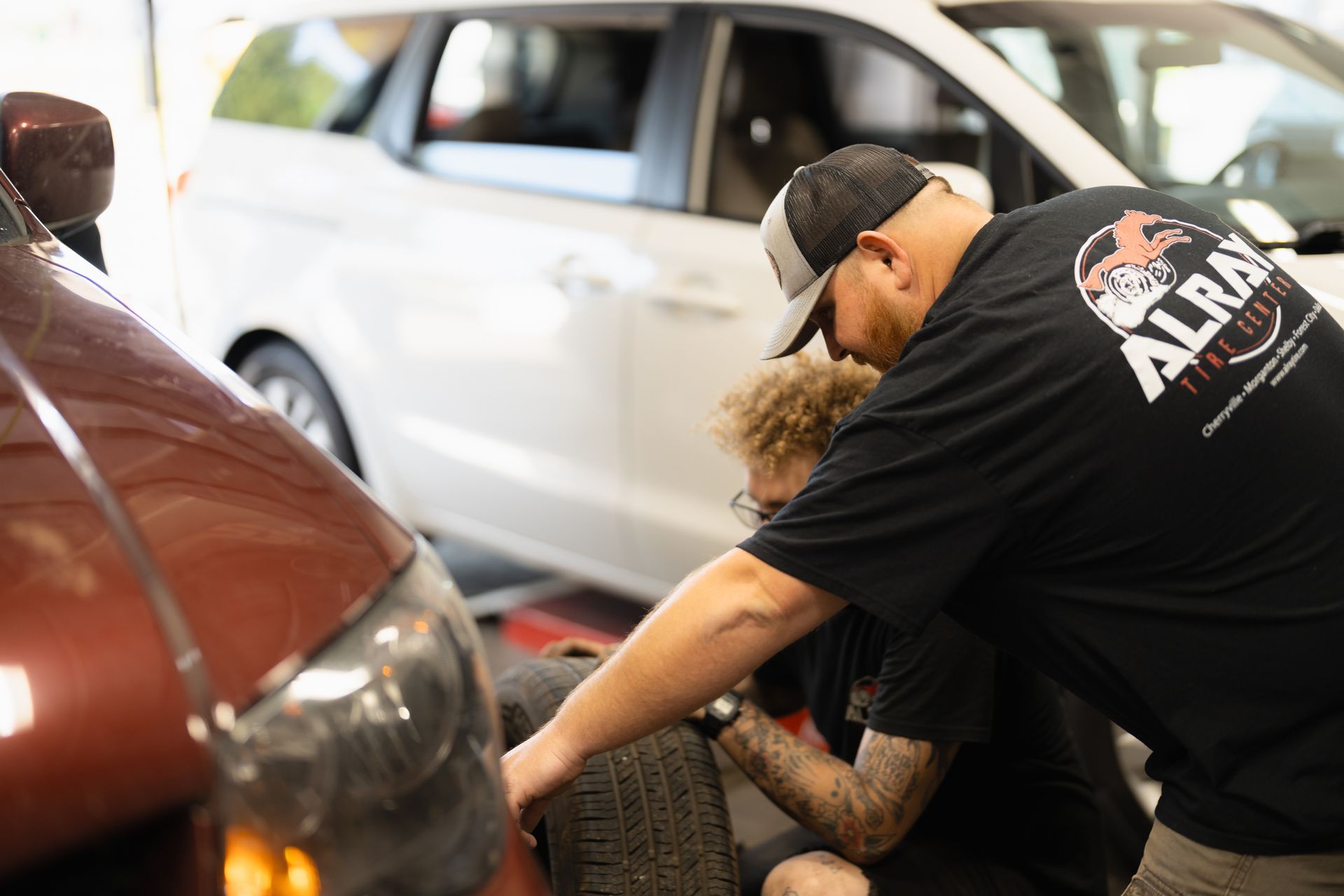 Mechanic repairing a car tire in a garage, with a white car in the background.