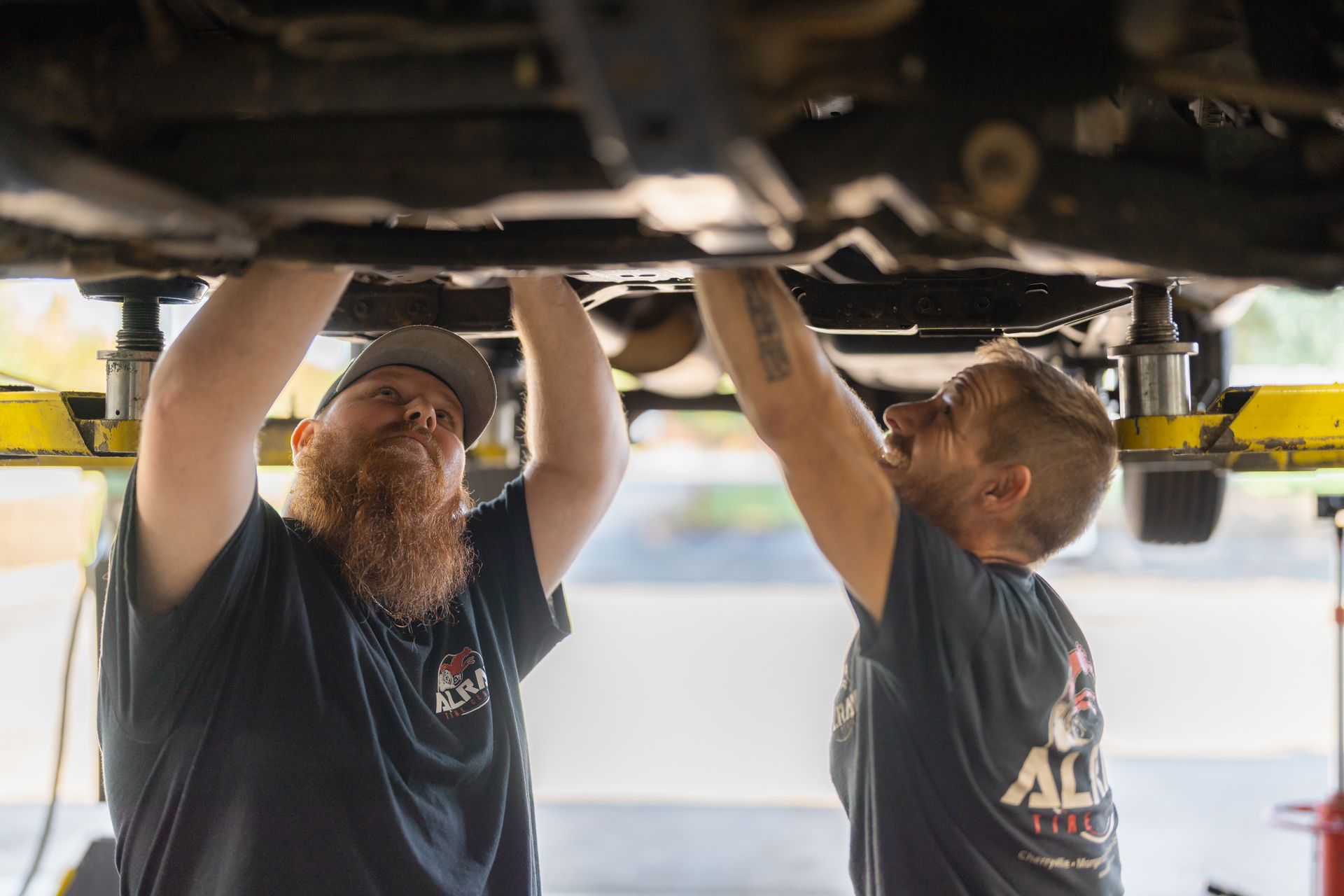 Two mechanics working under a lifted vehicle in a garage, using tools to inspect the underside.