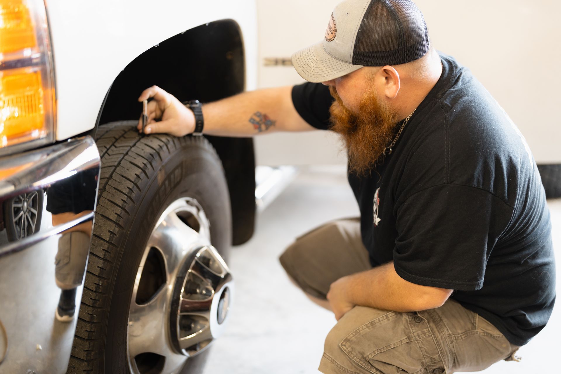 Mechanic crouching beside a truck, inspecting the front tire with a tool in a garage